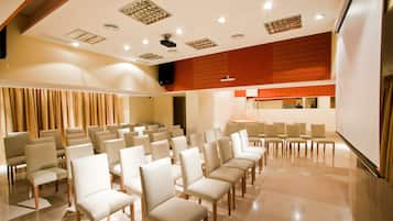 A conference room with rows of white chairs, a wooden ceiling, and a large screen.