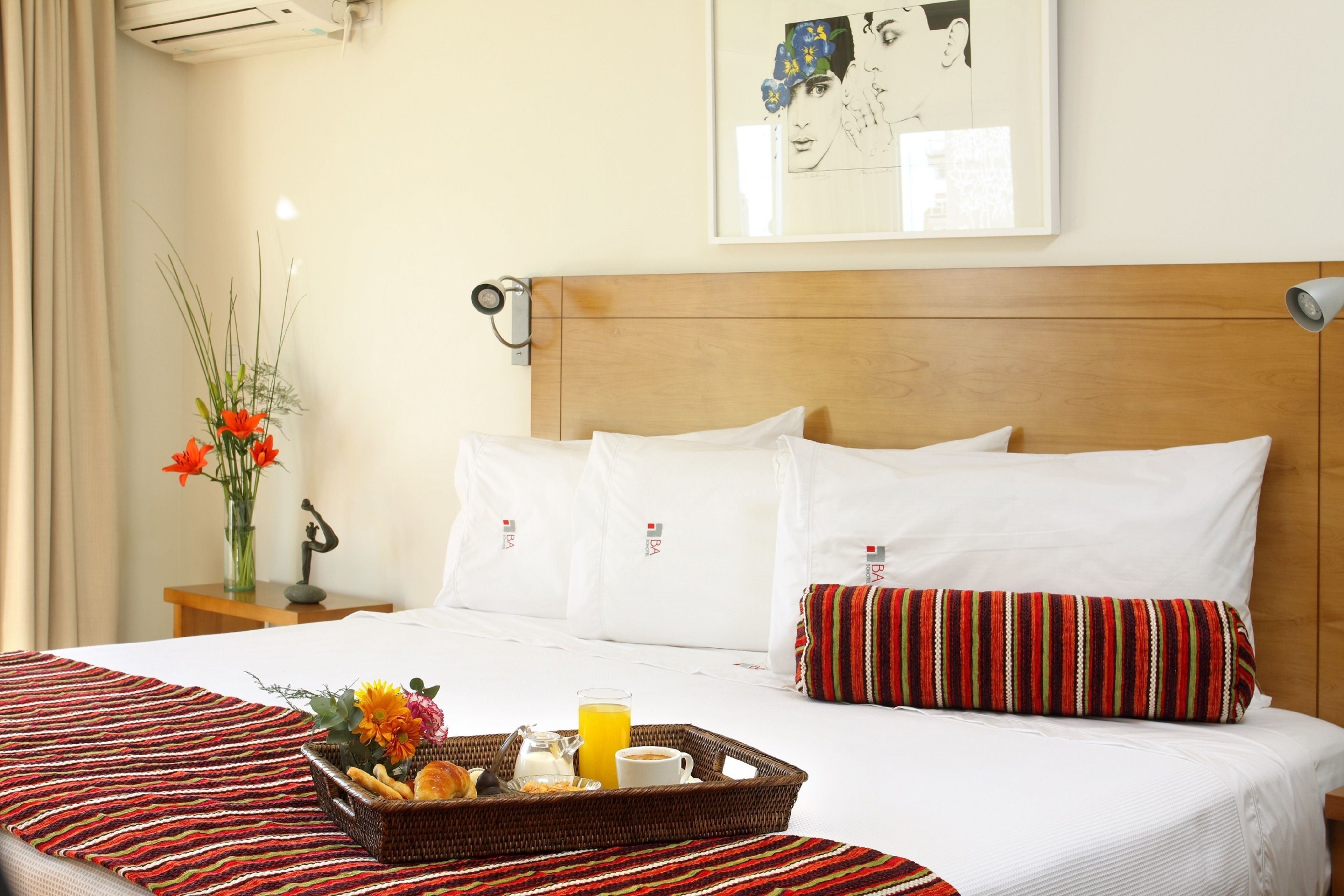 A hotel room with a neatly made bed, a breakfast tray on the bed, and a framed artwork on the wall.