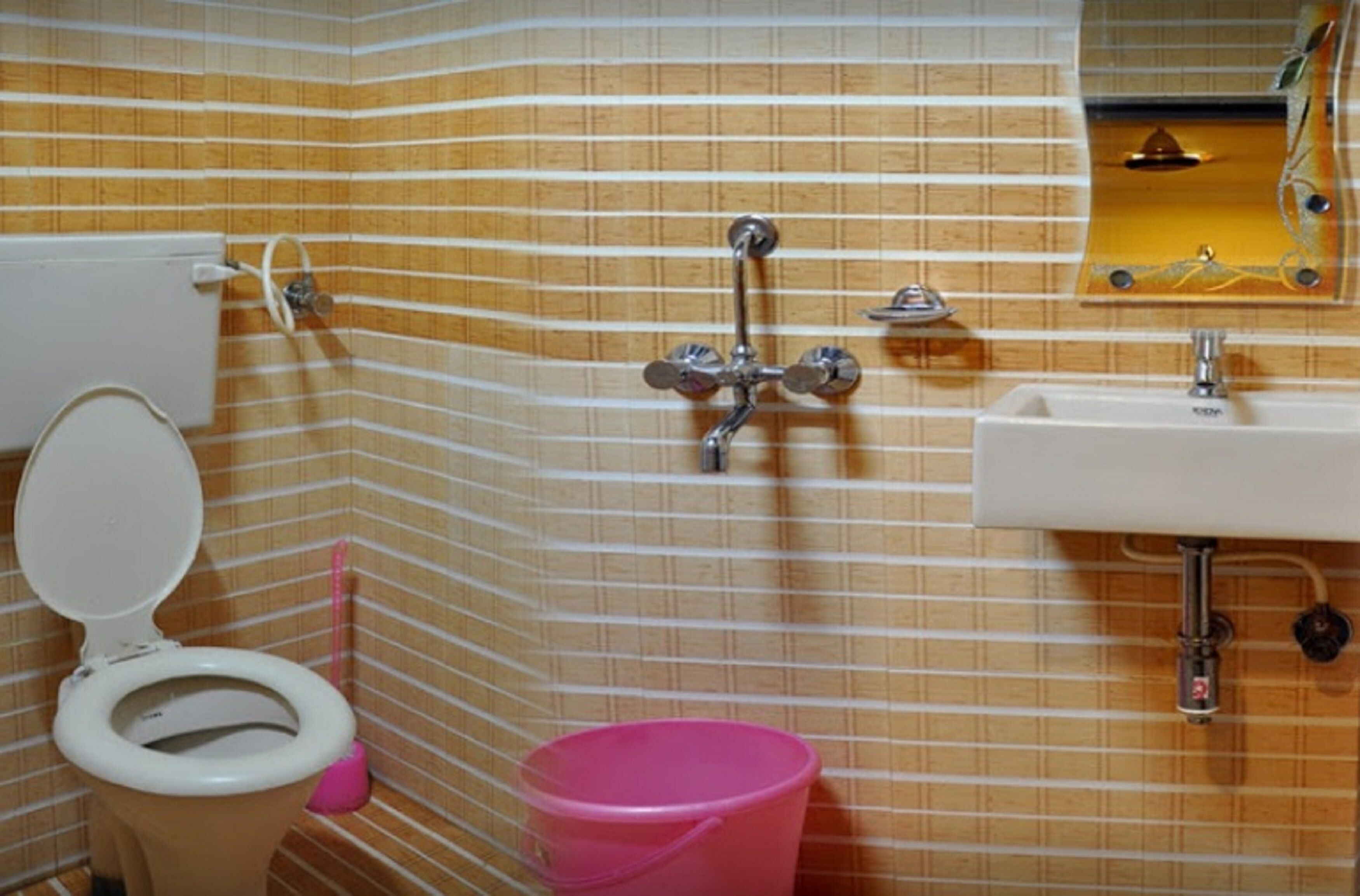 A bathroom with striped tile walls, a toilet, a pink bucket, and a sink with a mirror above it.