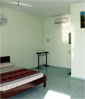 A bedroom with a bed, a standing fan, an air conditioning unit, and a small table.