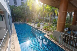 A swimming pool with clear blue tiles, surrounded by palm trees and plants, and a covered area with columns. - Honey Resort (Karon)