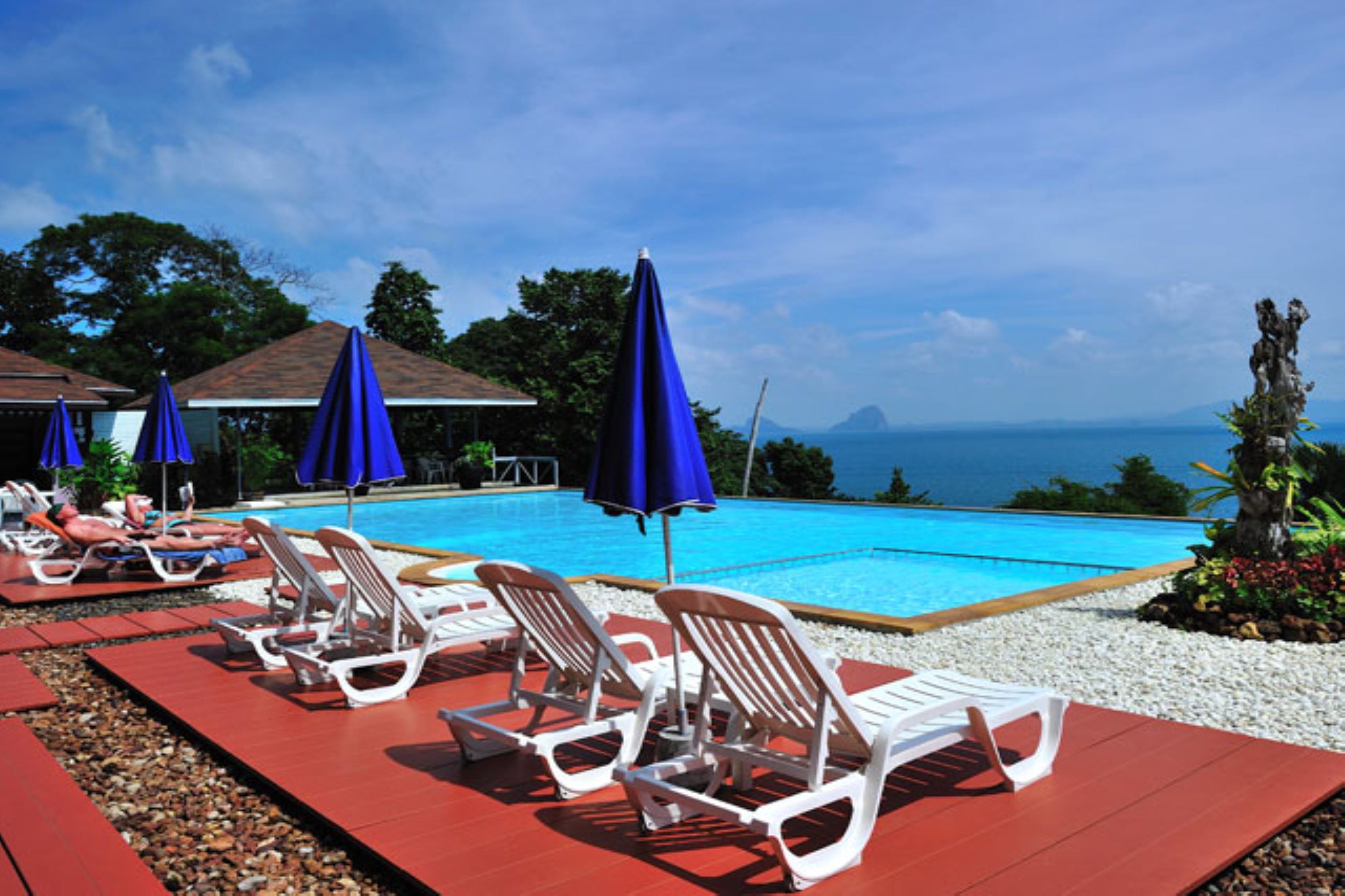 A poolside area with lounge chairs and umbrellas, a clear view of the ocean, and a tropical setting.