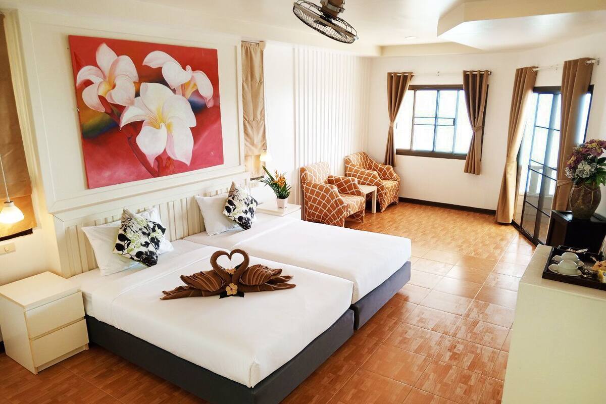 Une chambre d’hôtel avec un grand lit, deux tables de chevet, une commode, un tableau, une chaise, un bureau et une fenêtre avec des rideaux.