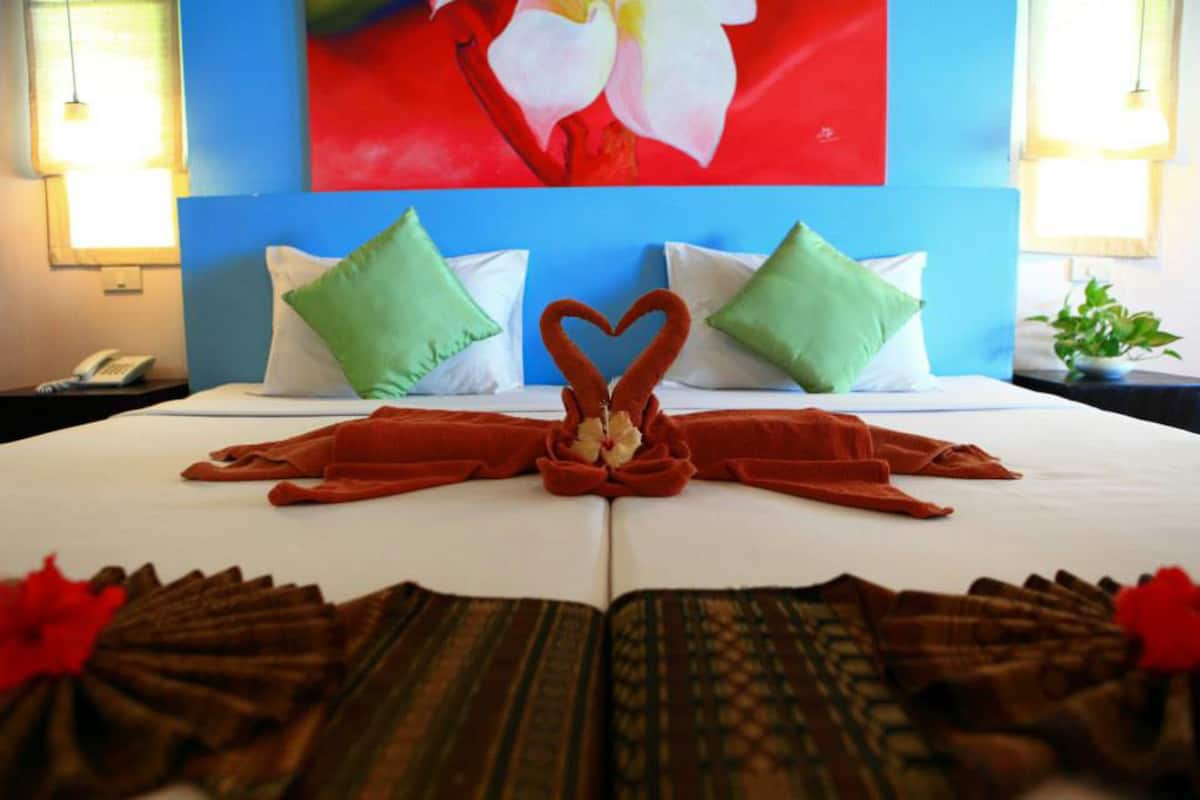Un lit bien fait, agrémenté d’une décoration en forme de cœur réalisée avec des serviettes, de coussins verts et d’une fleur rouge.