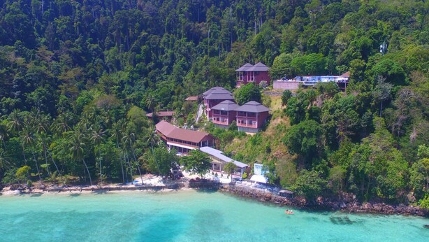 Een kustresort met meerdere gebouwen, waaronder een groot huis met een rood dak en enkele kleinere constructies, omringd door weelderige begroeiing en helder turquoise water.