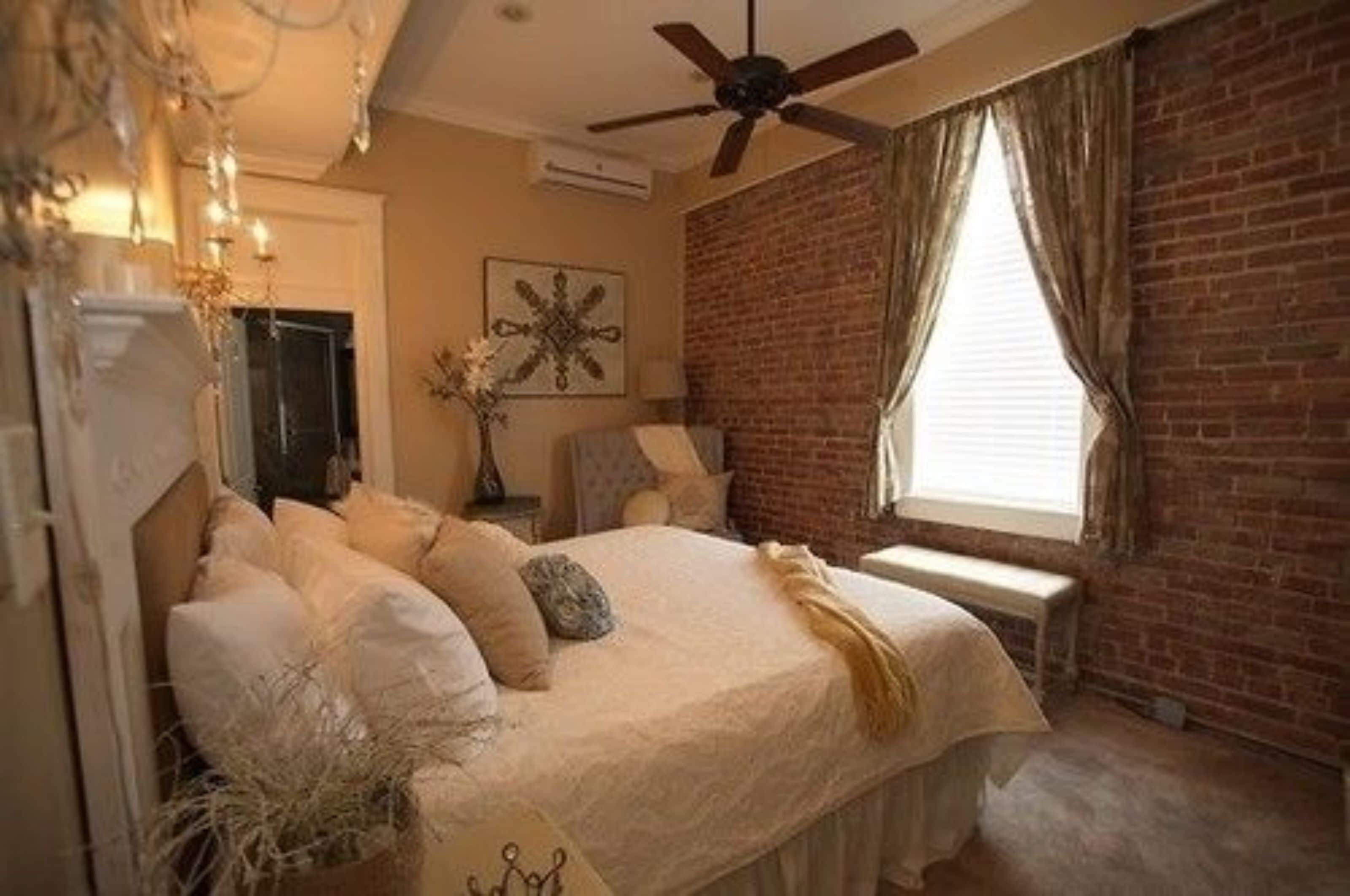 Un dormitorio con cama, ventilador de techo, pared de ladrillo y ventana con cortinas.