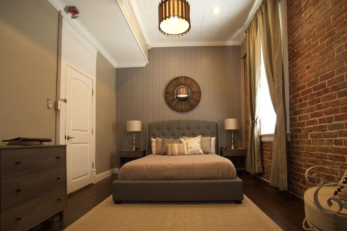 A bedroom with a large bed, a dresser, a nightstand, a wall clock, and a brick wall.