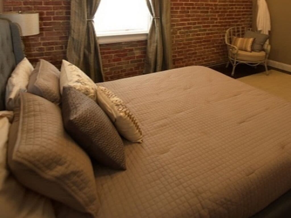 Una cama bien hecha con cubrecama acolchada y varias almohadas, una pared de ladrillo, una ventana con cortinas y una silla con cojines.