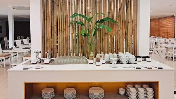 A modern dining area with a bamboo wall, neatly arranged white dining tables and chairs, and a buffet counter with plates and bowls. - Alba Hotel (Hue)