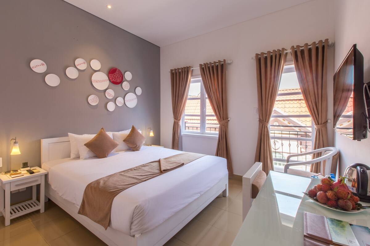 A hotel room with a bed, bedside table, and a desk with a fruit bowl.