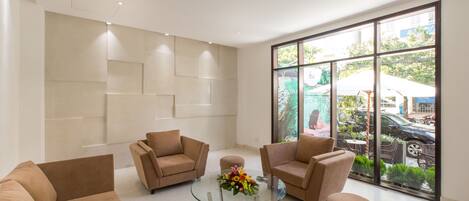 A modern lobby with beige sofas, a glass coffee table with a floral centerpiece, and large windows offering a view of the outdoors.