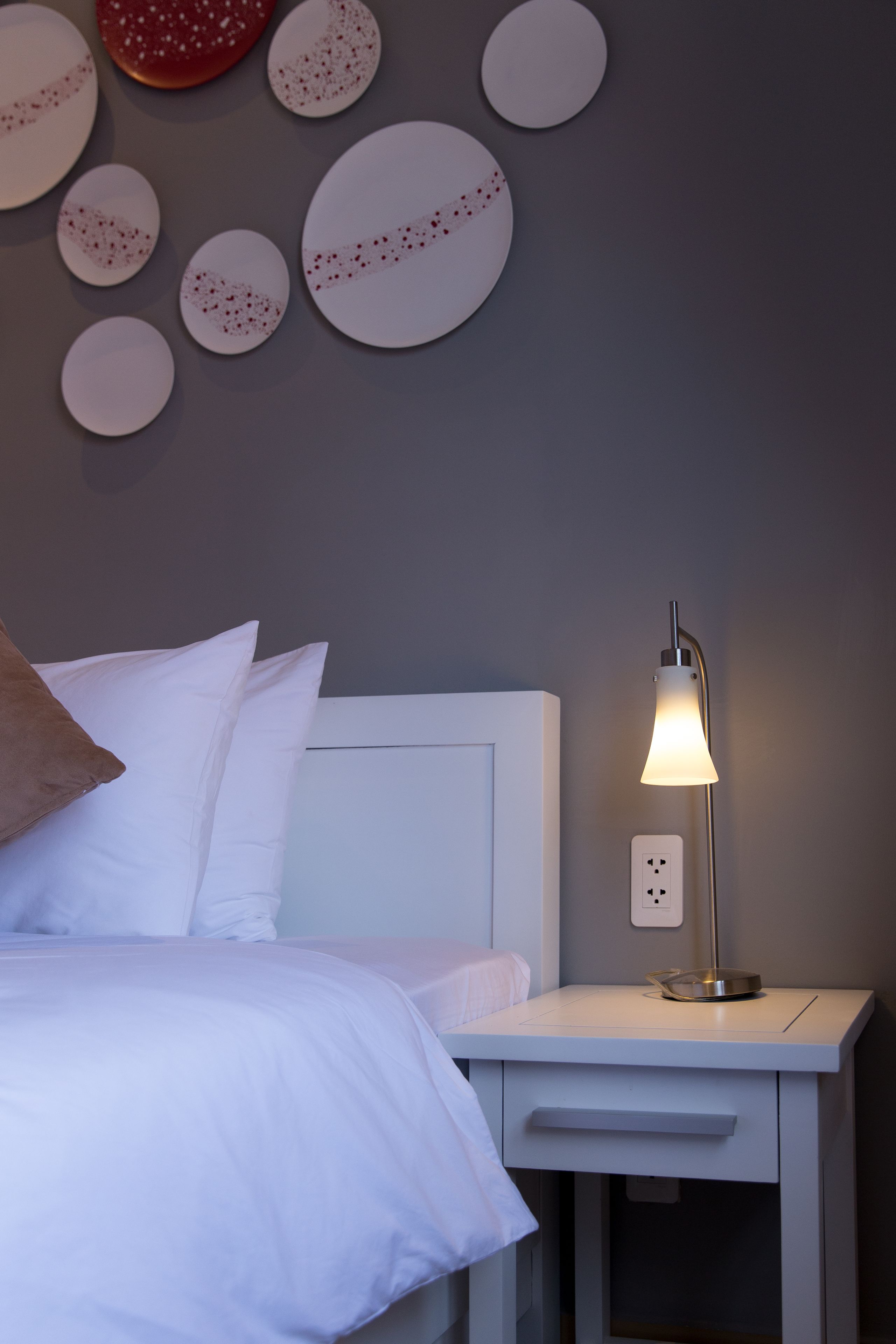 A neatly made bed with white linens, a bedside lamp, and a wall-mounted power outlet.