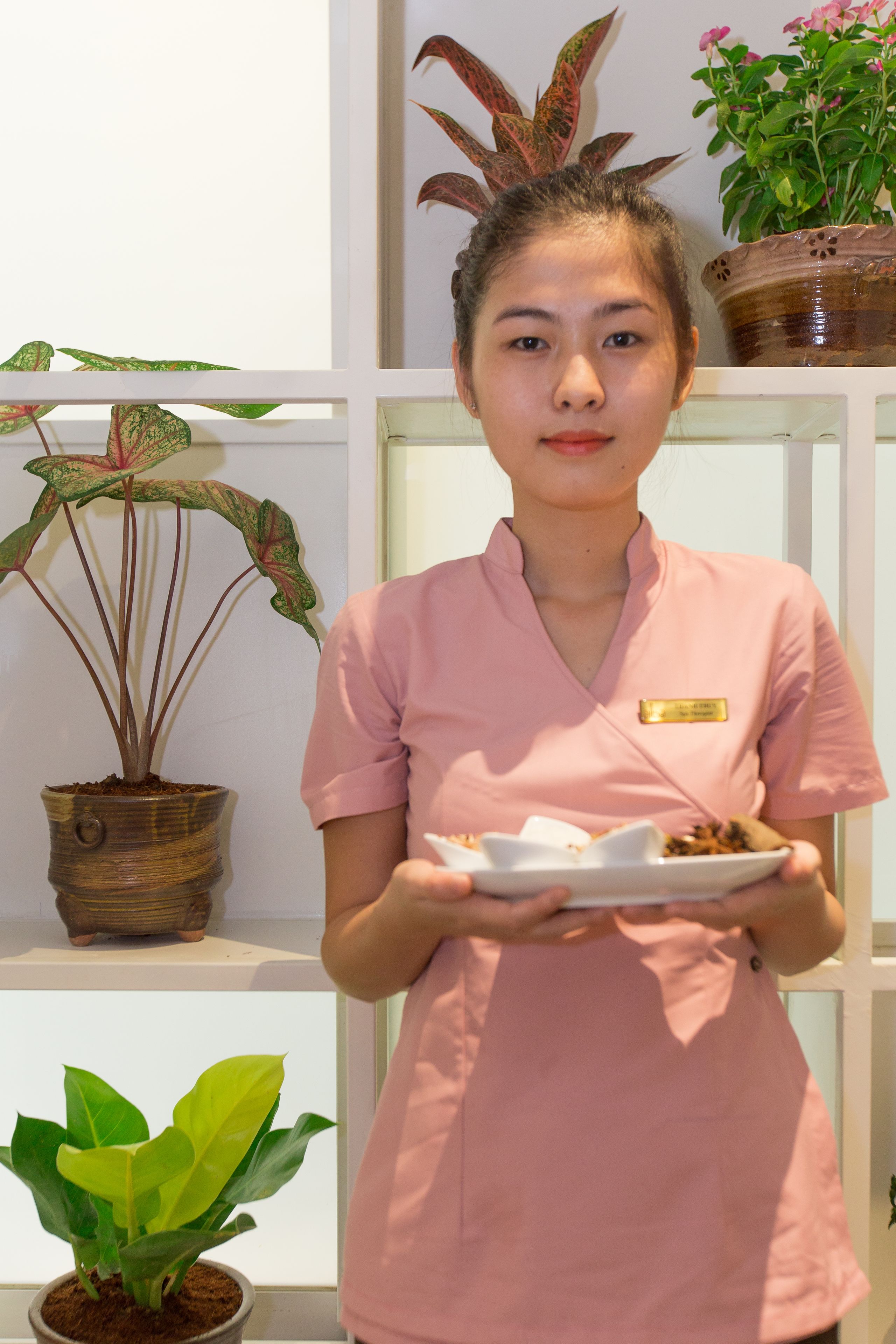 A person in a pink uniform holding a plate with food, standing in front of a shelf with potted plants.