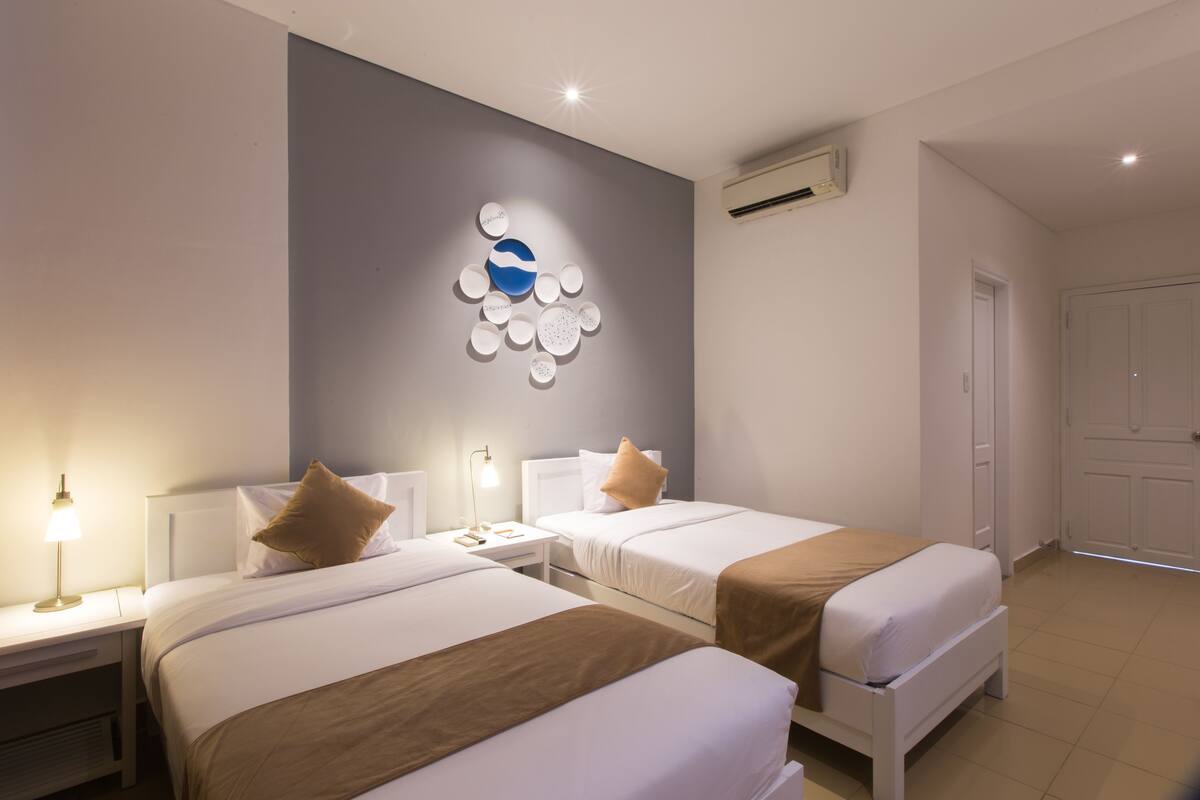 A hotel room with two beds, a wall-mounted artwork, a bedside table with a lamp, and a door.