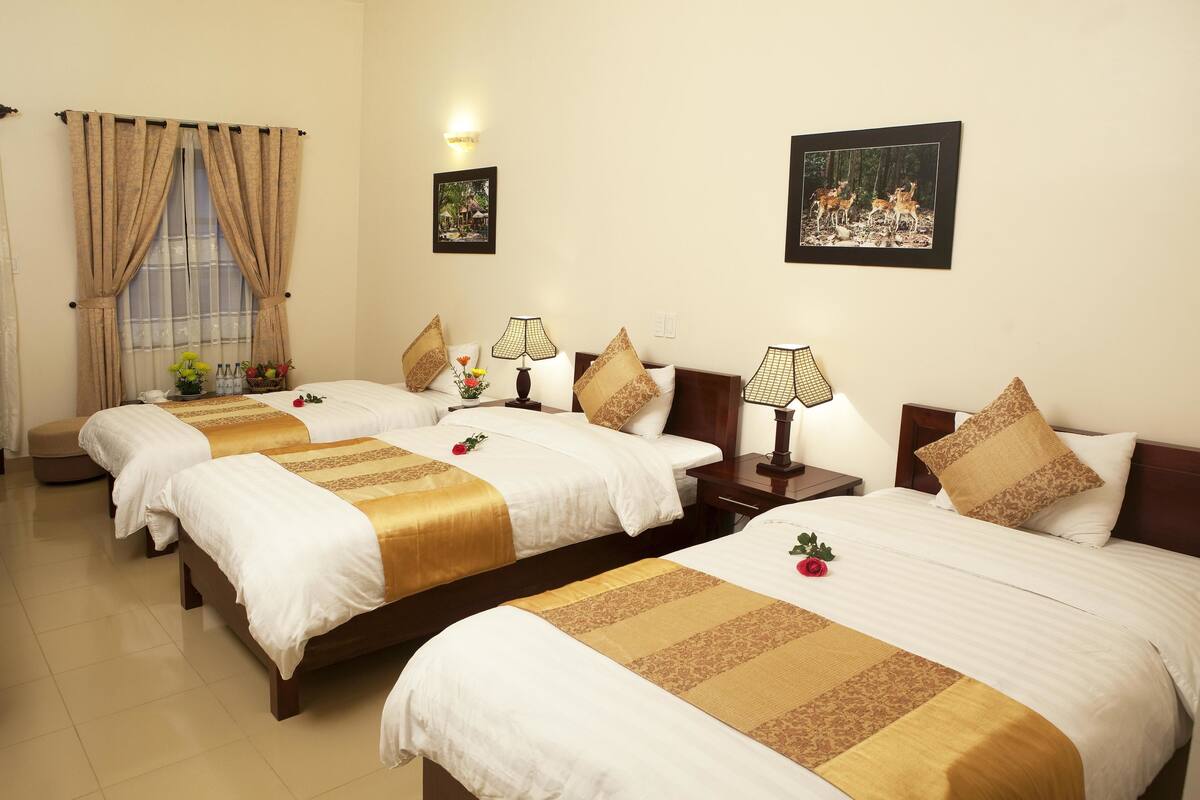 A hotel room with four beds, each with white bedding and gold accents, a window with beige curtains, and two wall-mounted lamps.