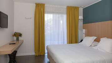 Habitación de hotel con una cama, una mesita de noche con un florero, un escritorio con una computadora y una ventana con cortinas amarillas.