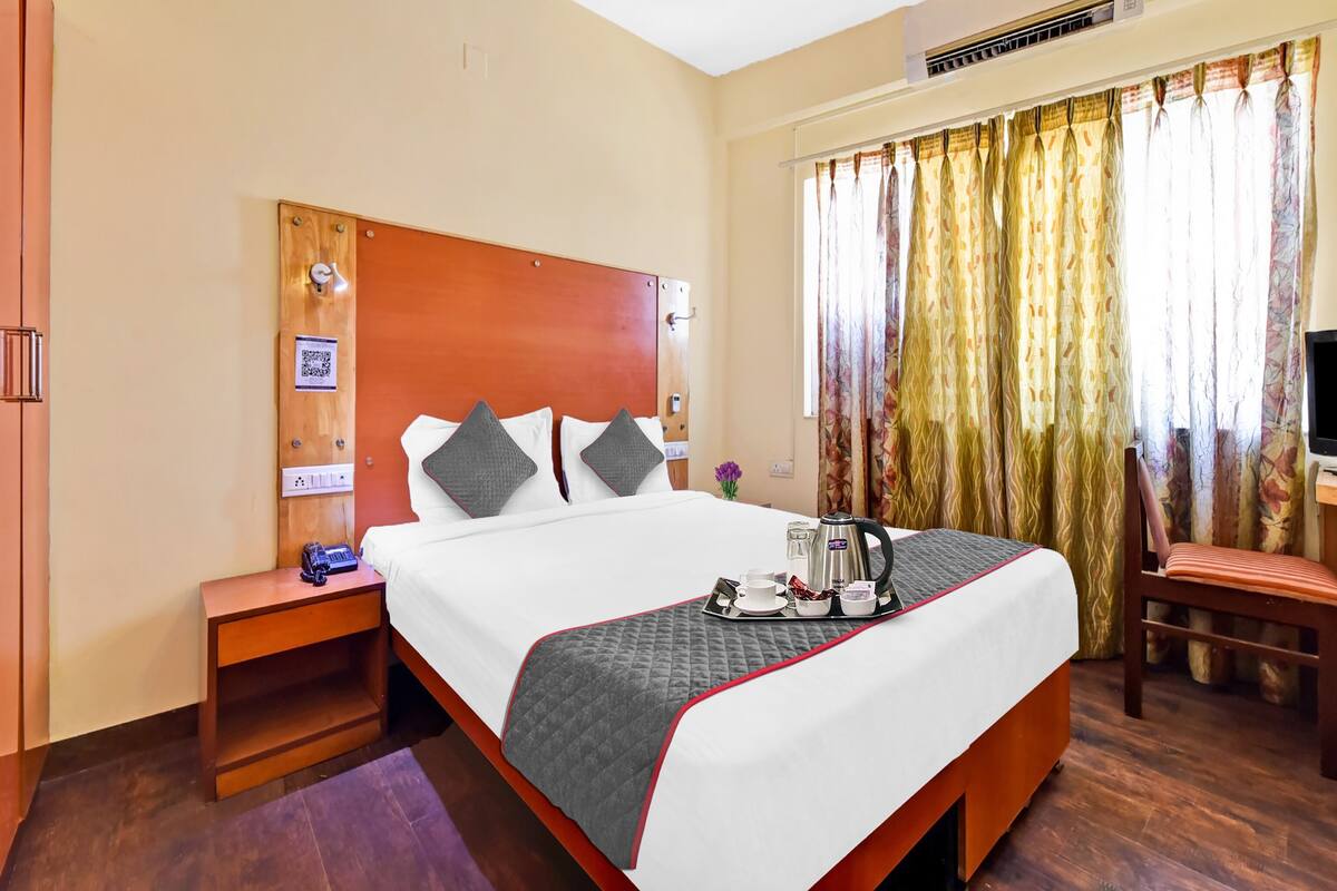 A hotel room with a large bed, a wooden headboard, a bedside table with a lamp, a chair, and a window with curtains.