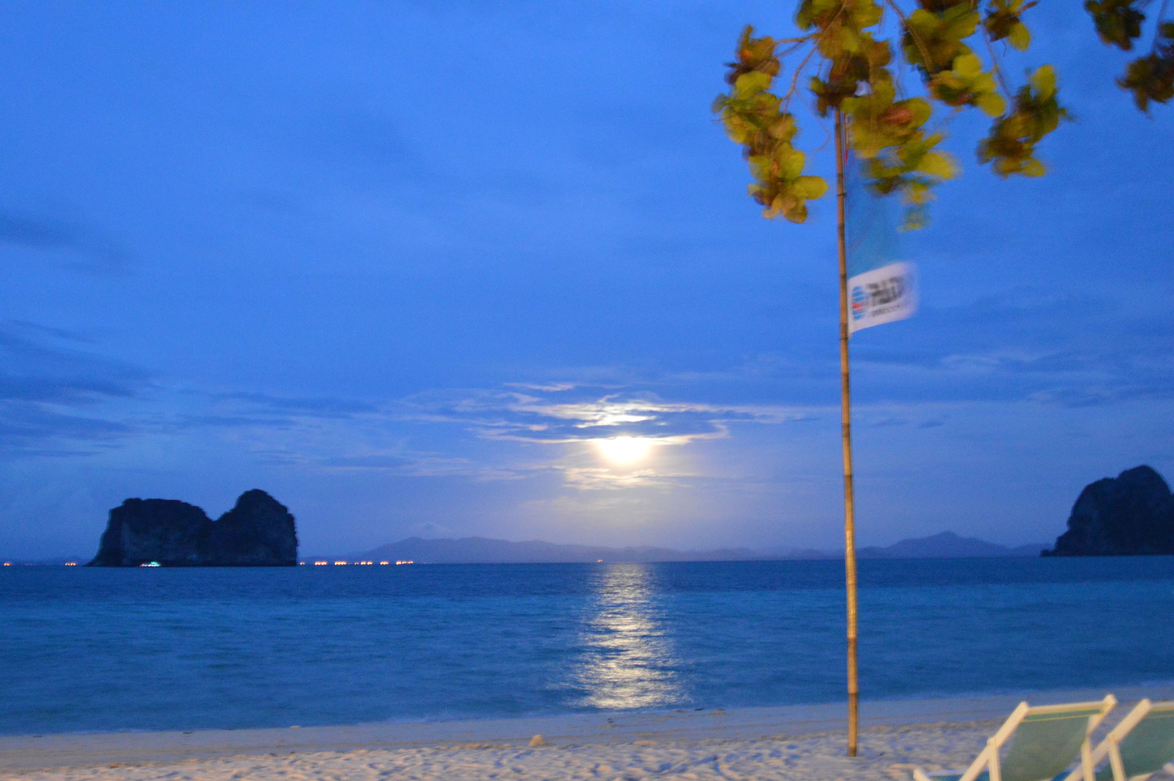 A beach scene with calm water, a visible moon, and distant rocky formations.