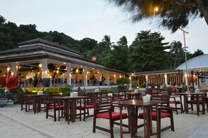 An outdoor restaurant with wooden tables and chairs, surrounded by greenery and palm trees.