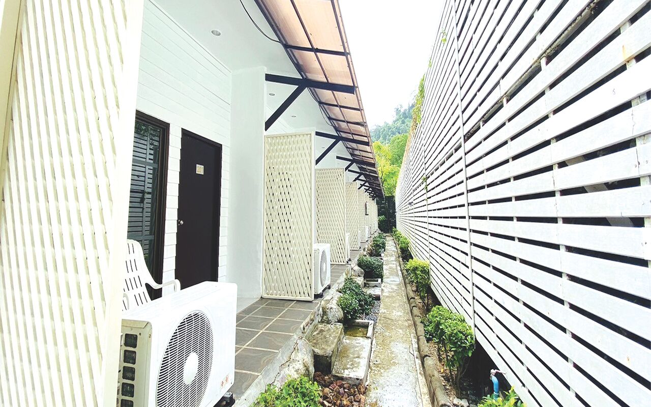 A narrow alleyway with a white building on one side and a wooden fence on the other. There are air conditioning units and a small garden area with plants and a water feature.