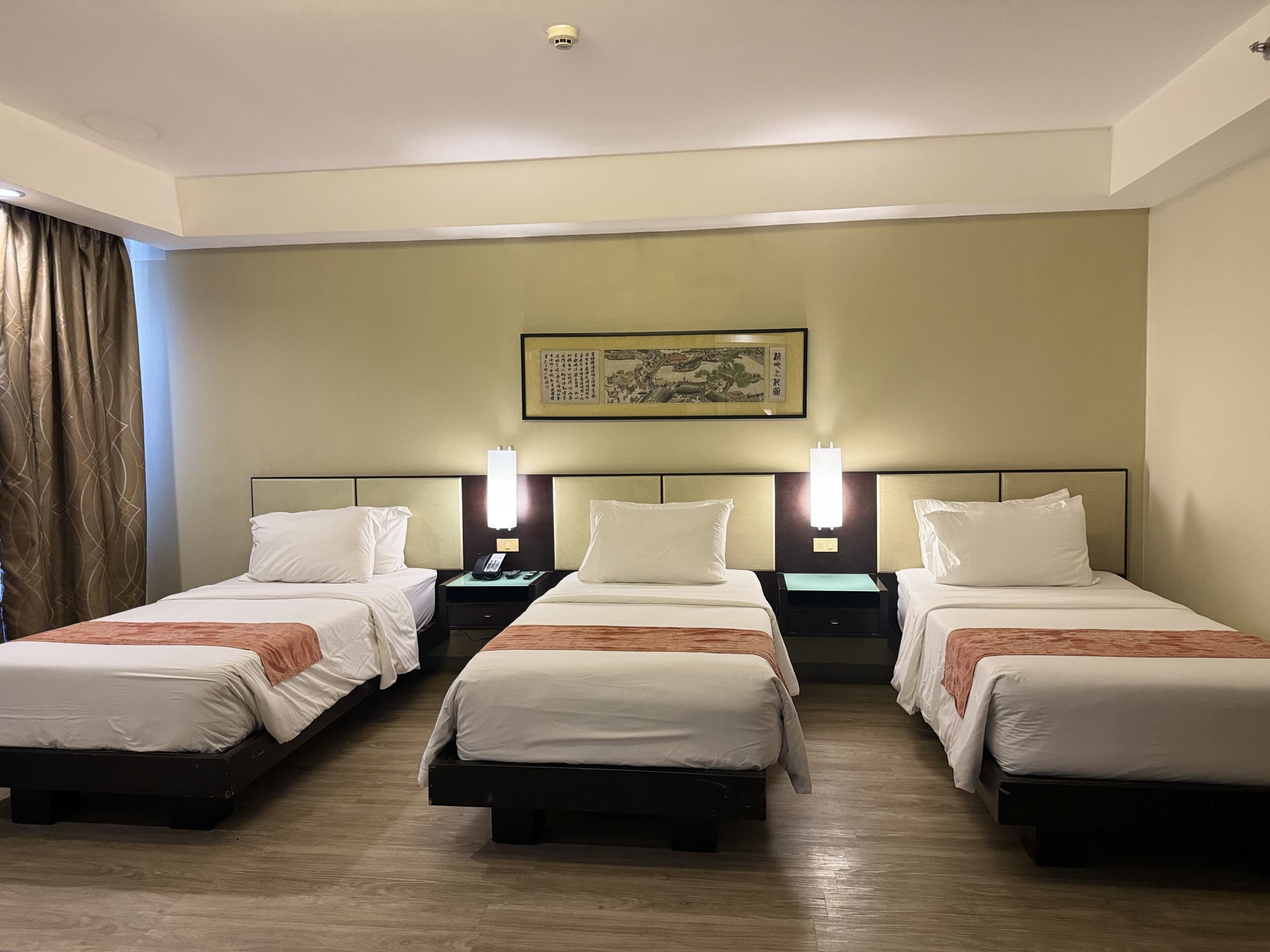 Habitación de hotel con tres camas, cada una con ropa blanca y una manta marrón. Hay mesitas de noche con lámparas y un teléfono en la pared. Encima de las camas, un cuadro enmarcado.