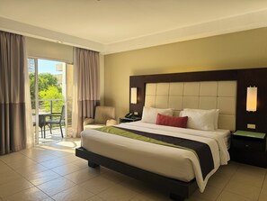 A hotel room with a large bed, a nightstand, a chair, and a view of greenery through the window. - Circle Inn - Iloilo City Center (Iloilo)