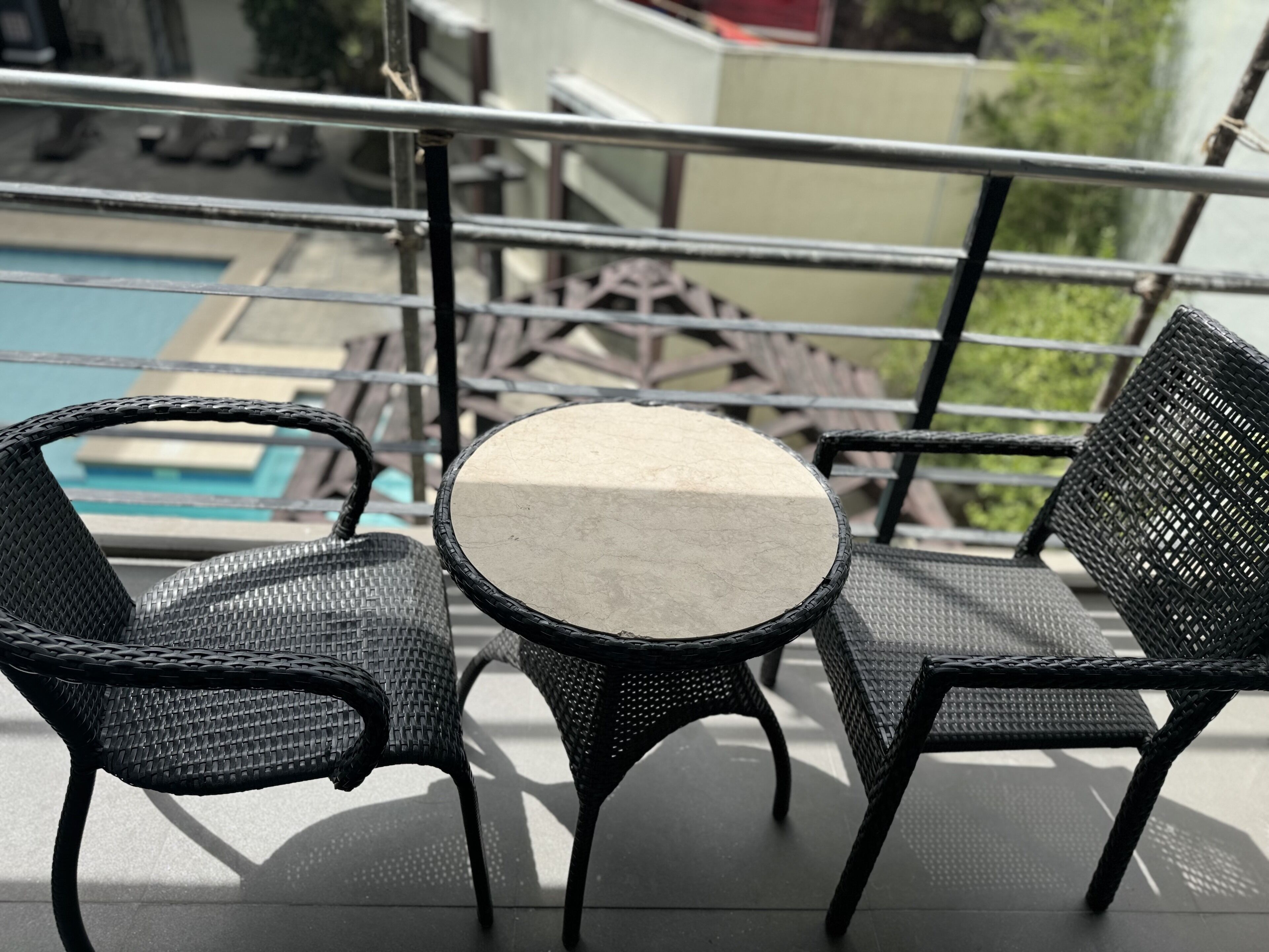 A balcony with a round table and two wicker chairs, overlooking a swimming pool.