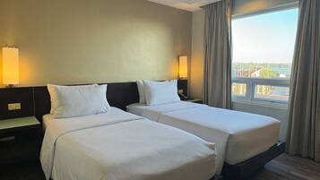A hotel room with two beds, a nightstand, a lamp, a window with curtains, and a view of the city.