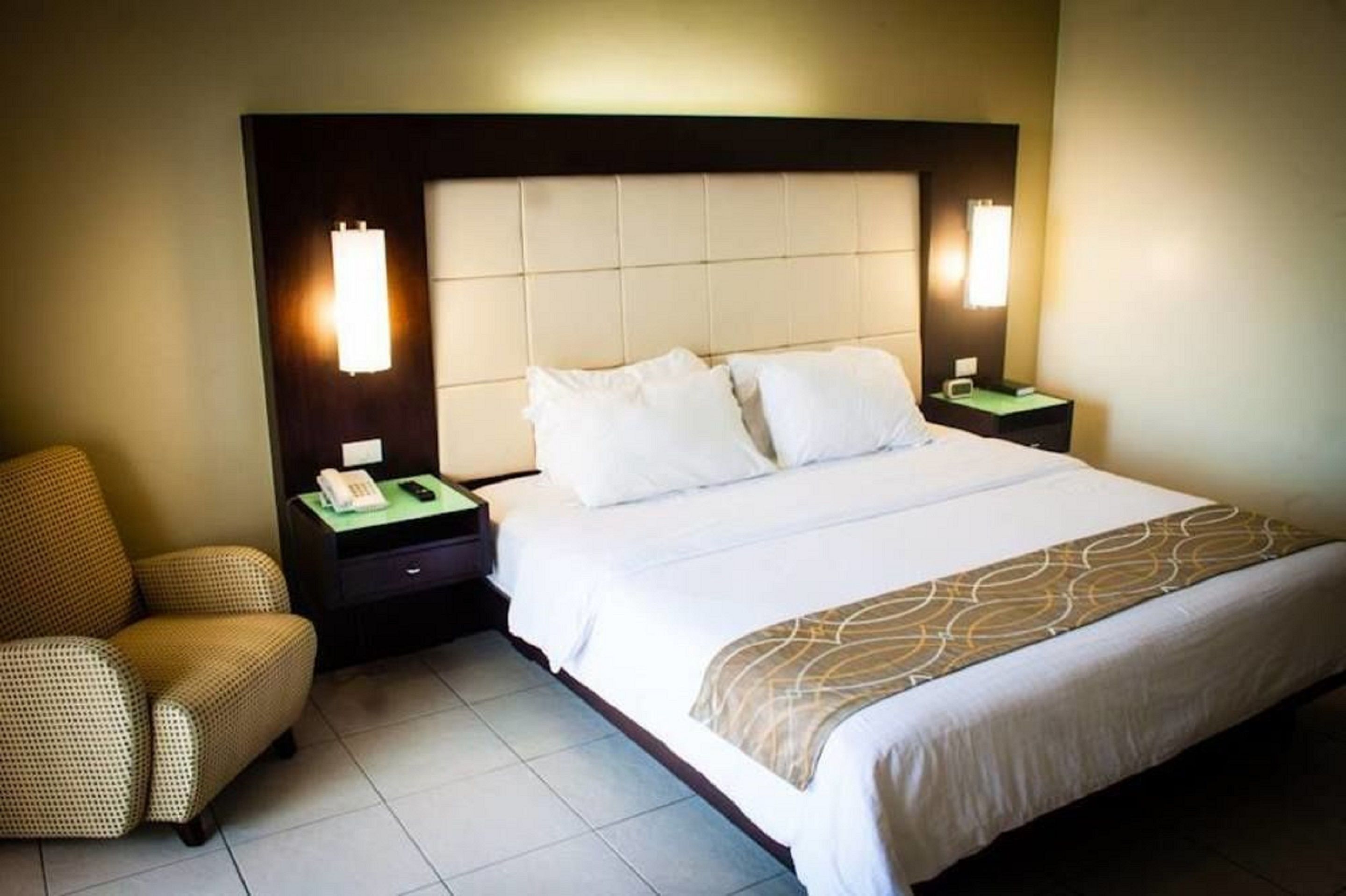 A hotel room with a large bed, two bedside tables, a chair, and a headboard with built-in lighting.
