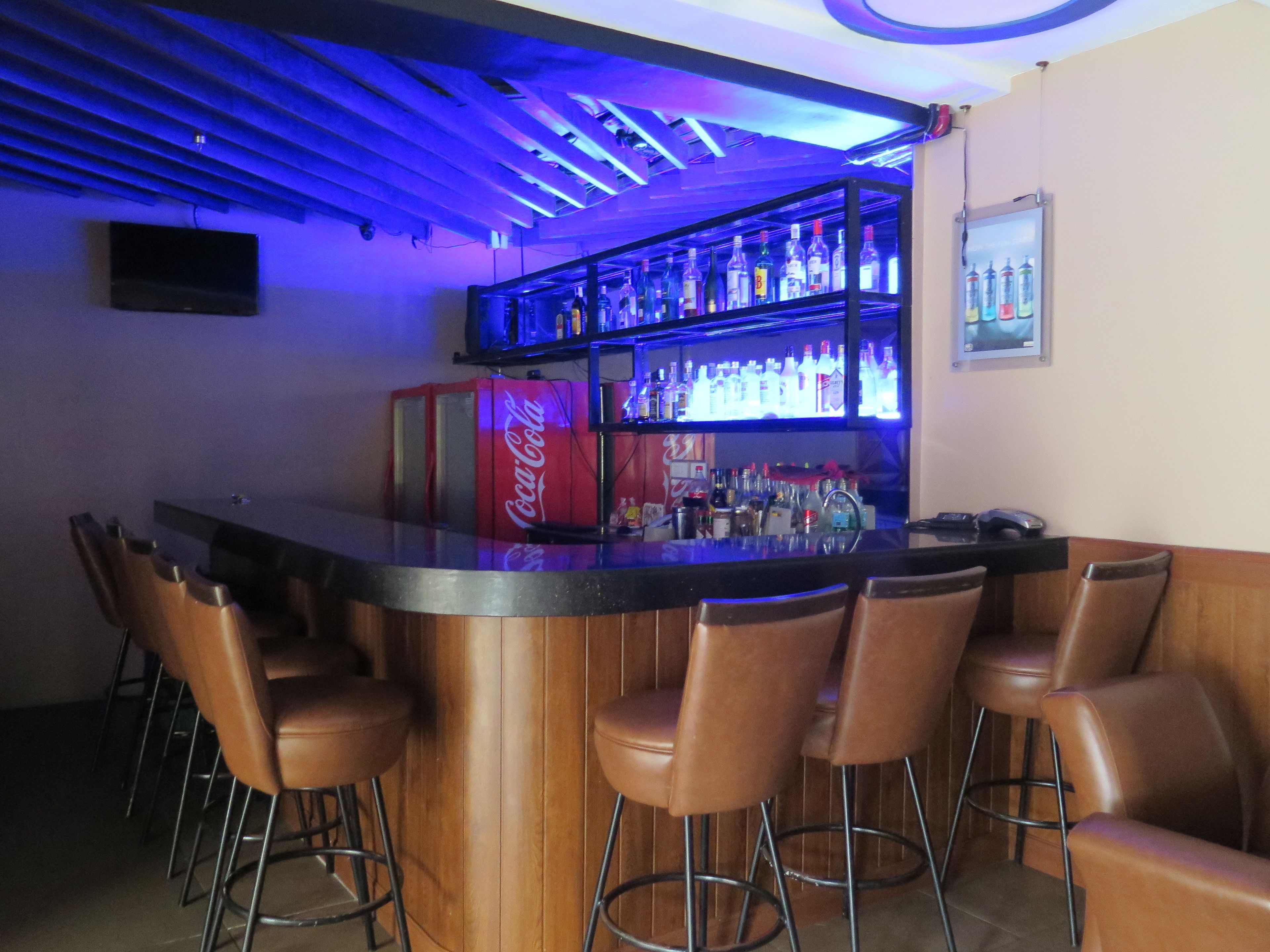 Un espace bar comprenant un comptoir, des tabourets de bar, des étagères remplies de bouteilles et un réfrigérateur pour les boissons, notamment les Coca-Cola.
