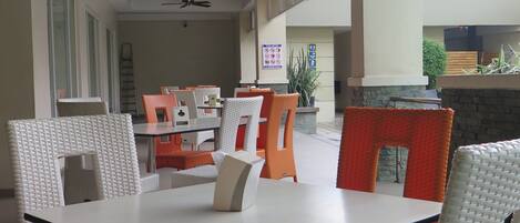Un amplio espacio de comedor al aire libre con sillas de mimbre blancas y naranjas, una mesa con cubierta de vidrio, ventiladores de techo y grandes ventanas que ofrecen luz natural.