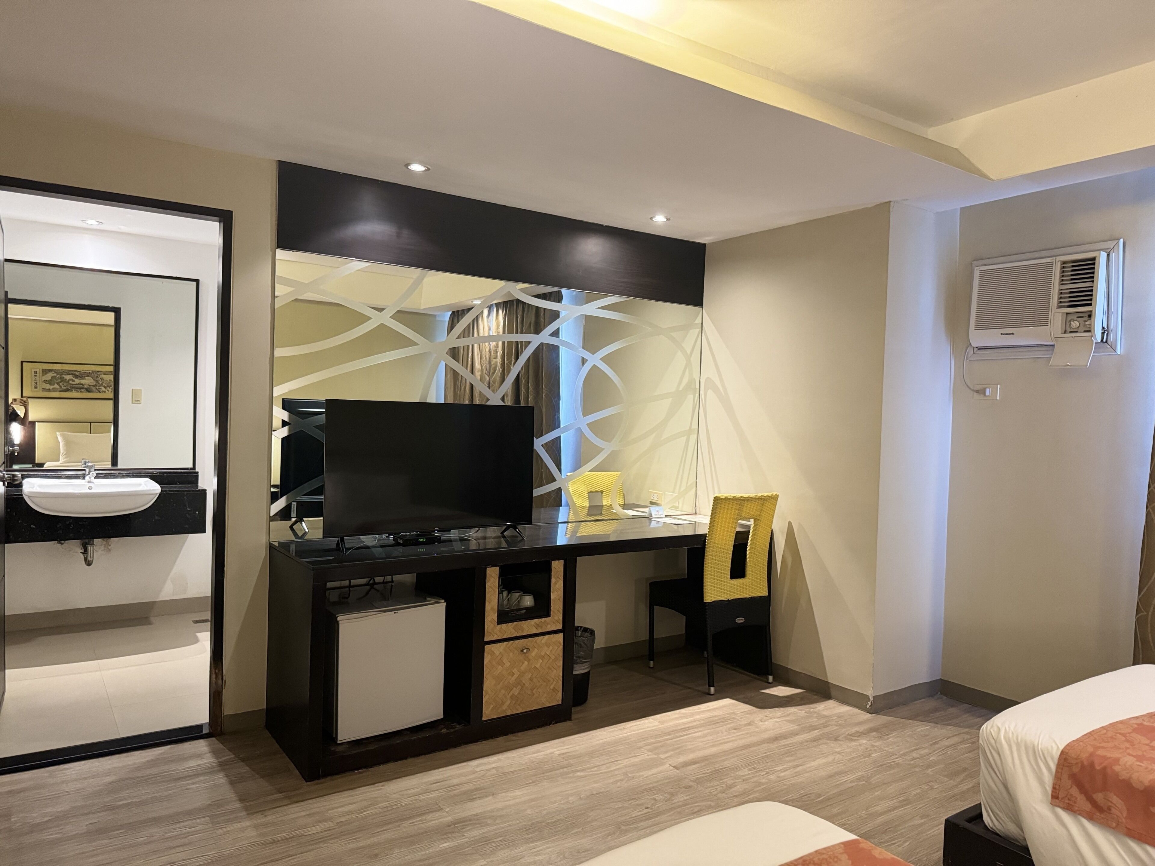 A hotel room with a bed, a desk with a TV, a small fridge, a chair, and a bathroom with a sink and mirror.