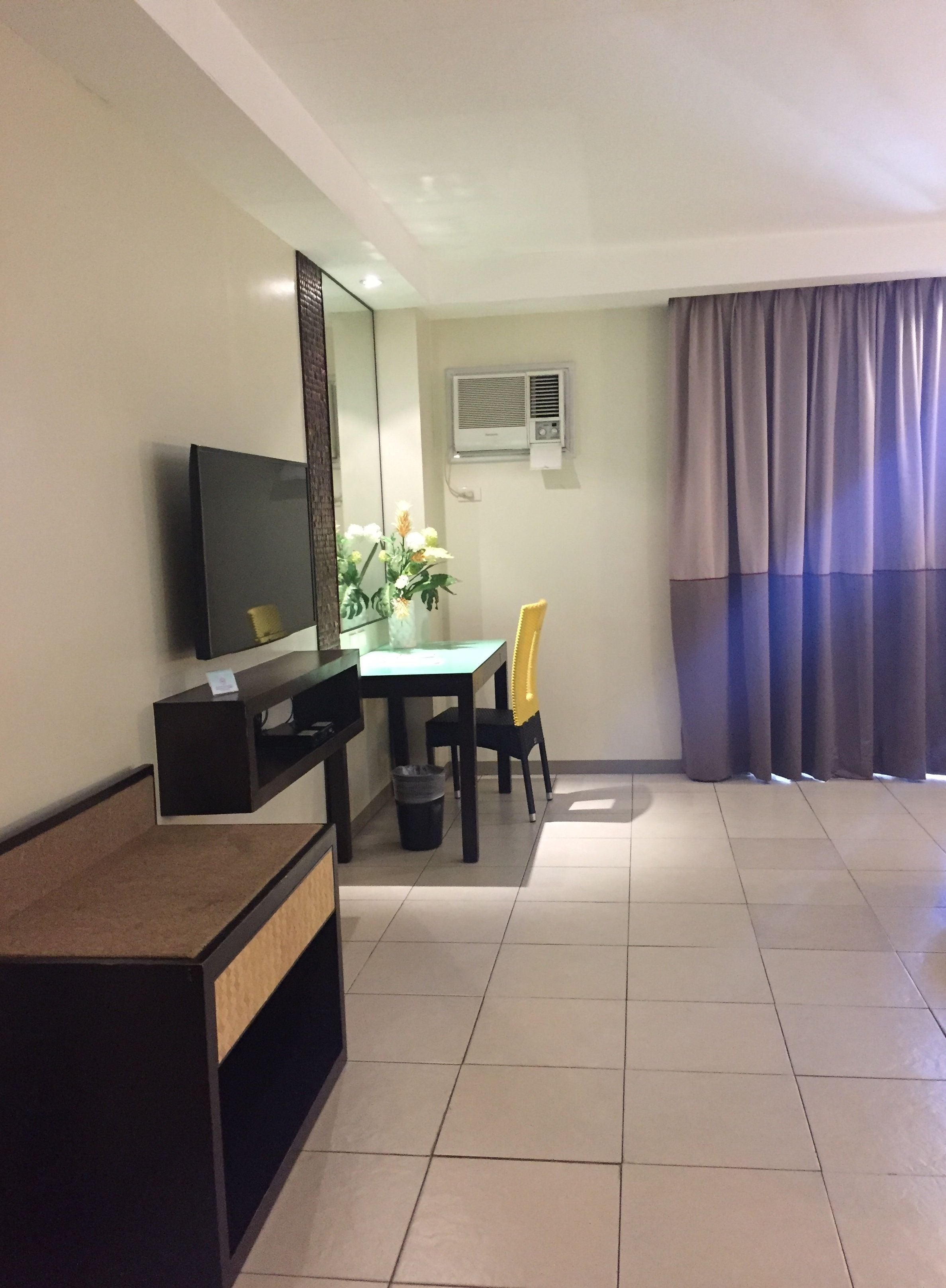 Habitación de hotel con televisor de pantalla plana, una mesita con una silla, una planta en maceta y una ventana con cortinas.