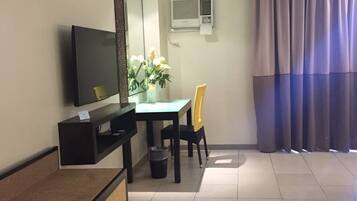 Habitación de hotel con televisor de pantalla plana, una mesita con una silla, una planta en maceta y una ventana con cortinas.