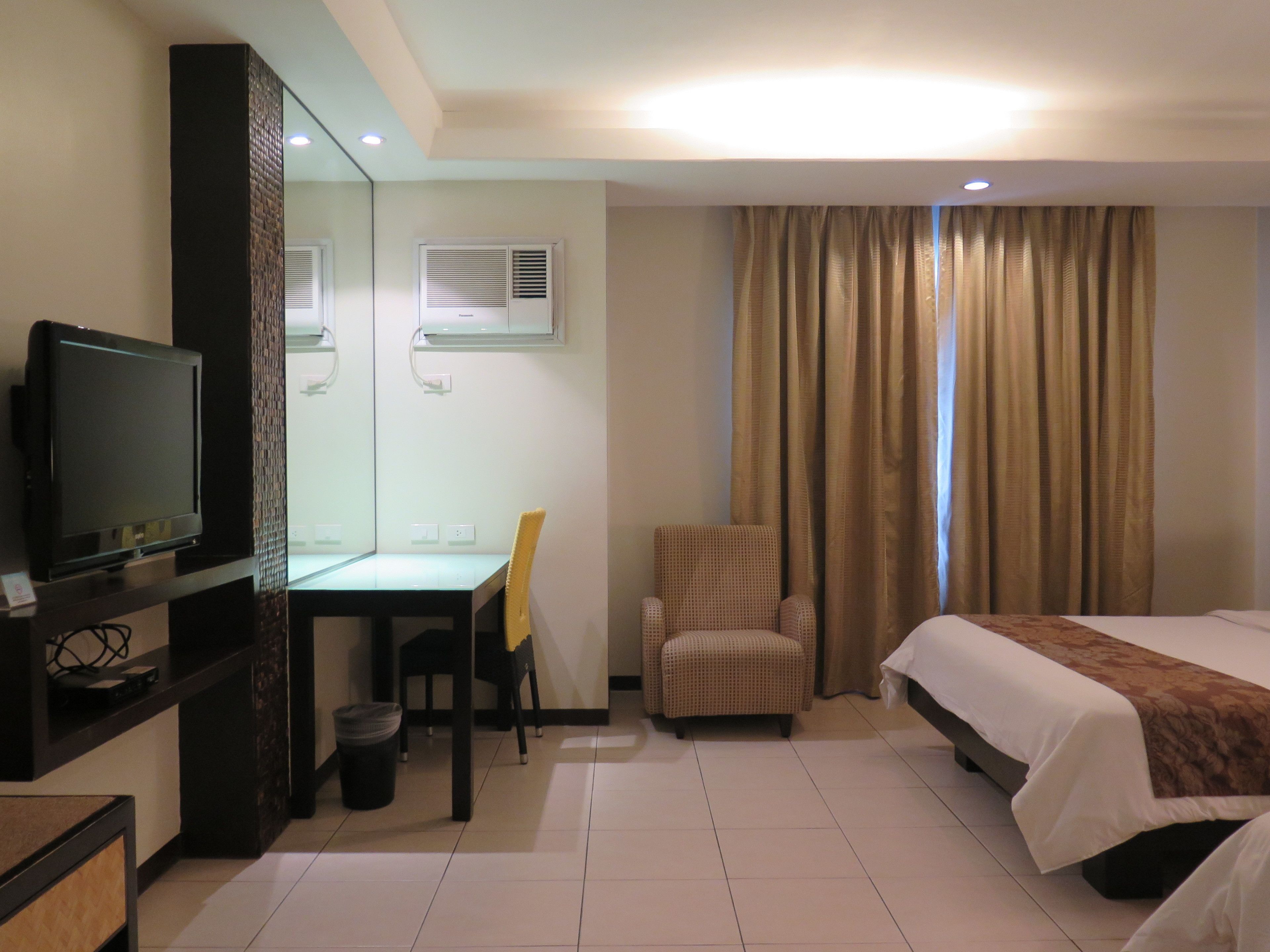 A hotel room with a bed, a desk with a chair, a television, and an air conditioning unit.