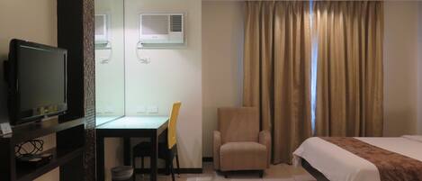 A hotel room with a bed, a desk with a chair, a television, and an air conditioning unit.