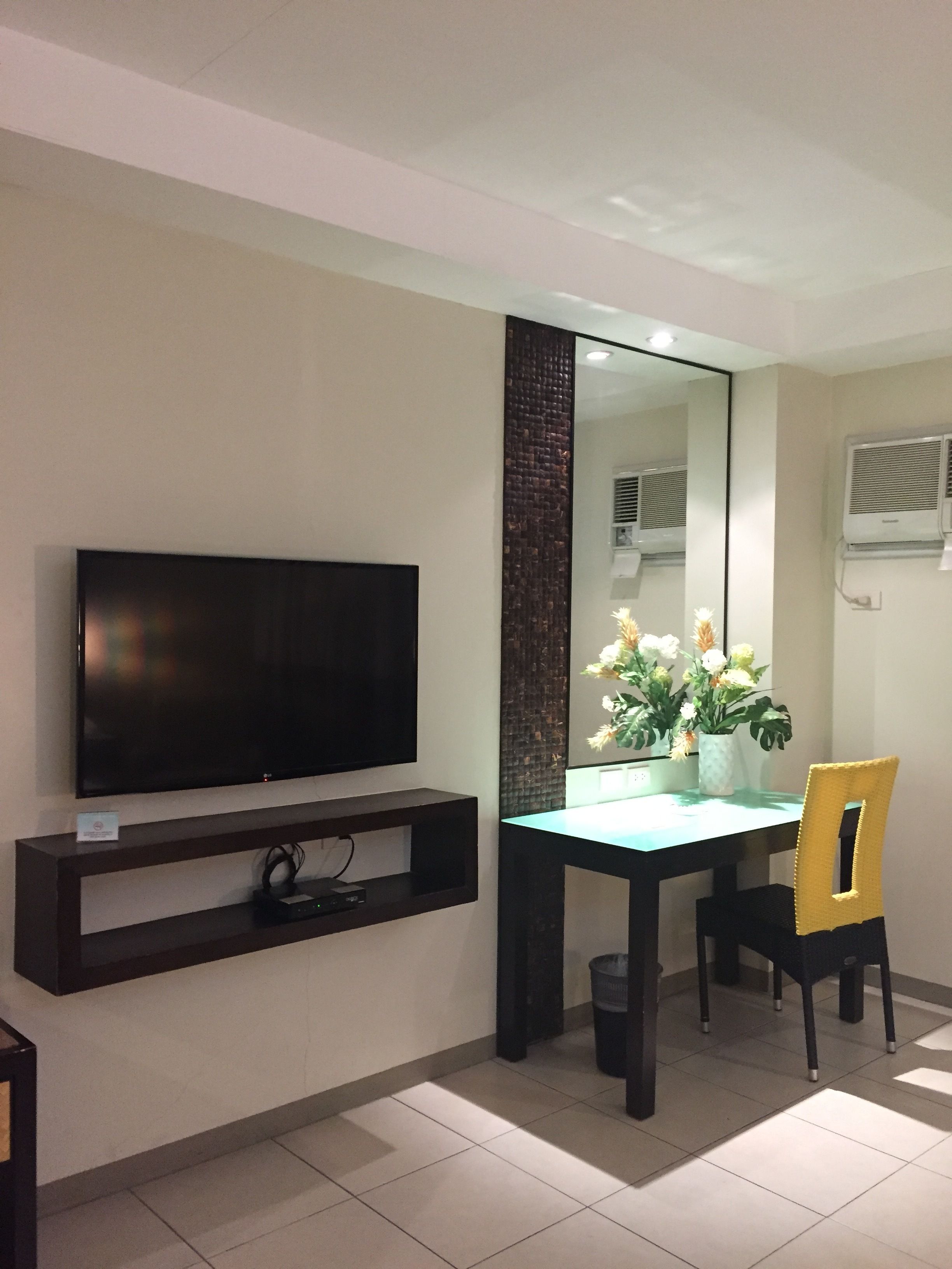 A modern hotel room with a flat-screen TV mounted on the wall, a dining table with chairs, and a large mirror reflecting the room.