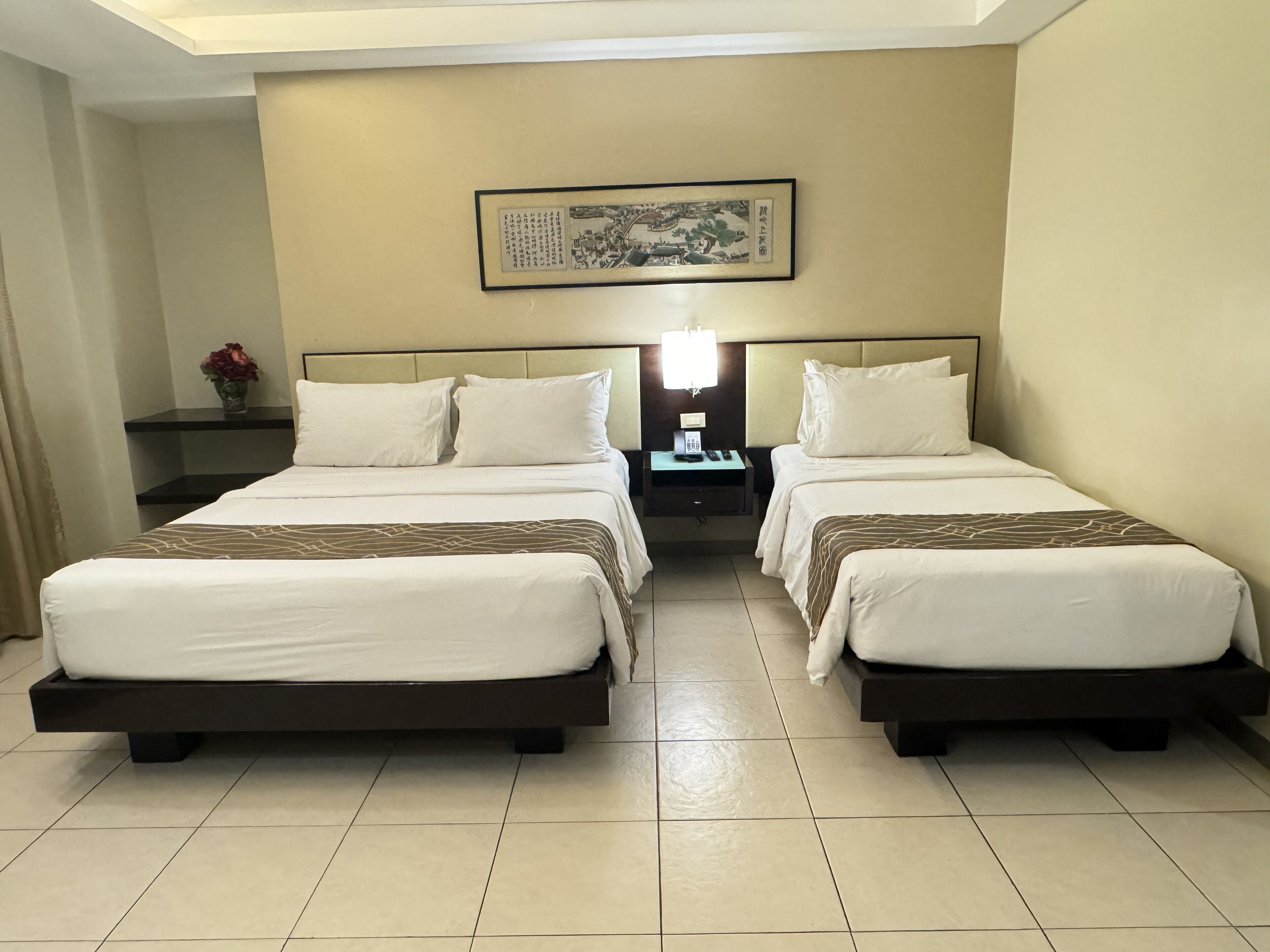 A hotel room with two beds, a nightstand with a lamp, a framed picture on the wall, and a vase with flowers on the nightstand.