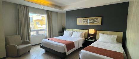 Habitación de hotel con dos camas, una silla, una mesita de noche con lámpara, una ventana con cortinas y una obra de arte colgada en la pared.