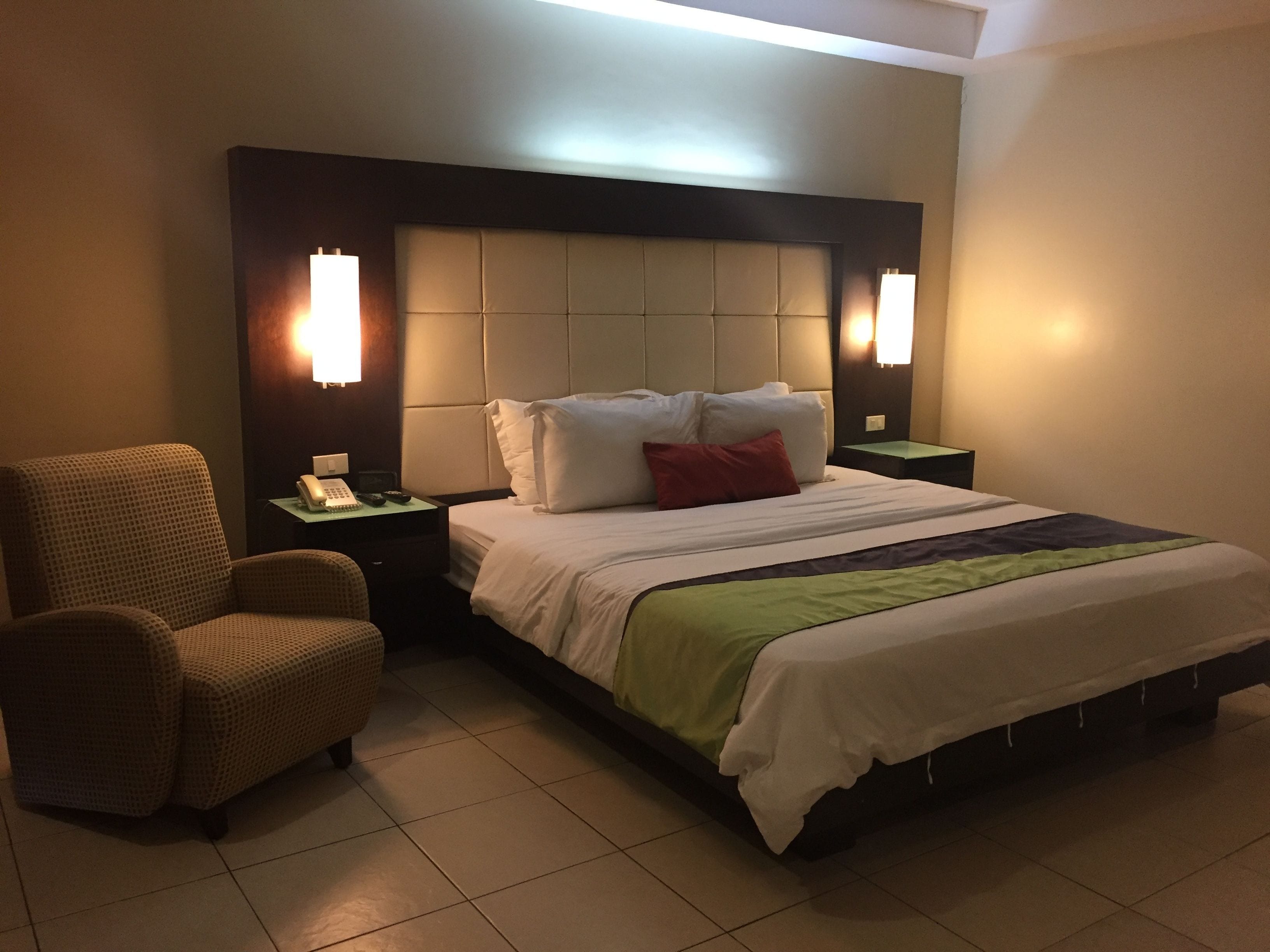 A hotel room with a bed, a chair, a nightstand with a phone, and wall-mounted lights.