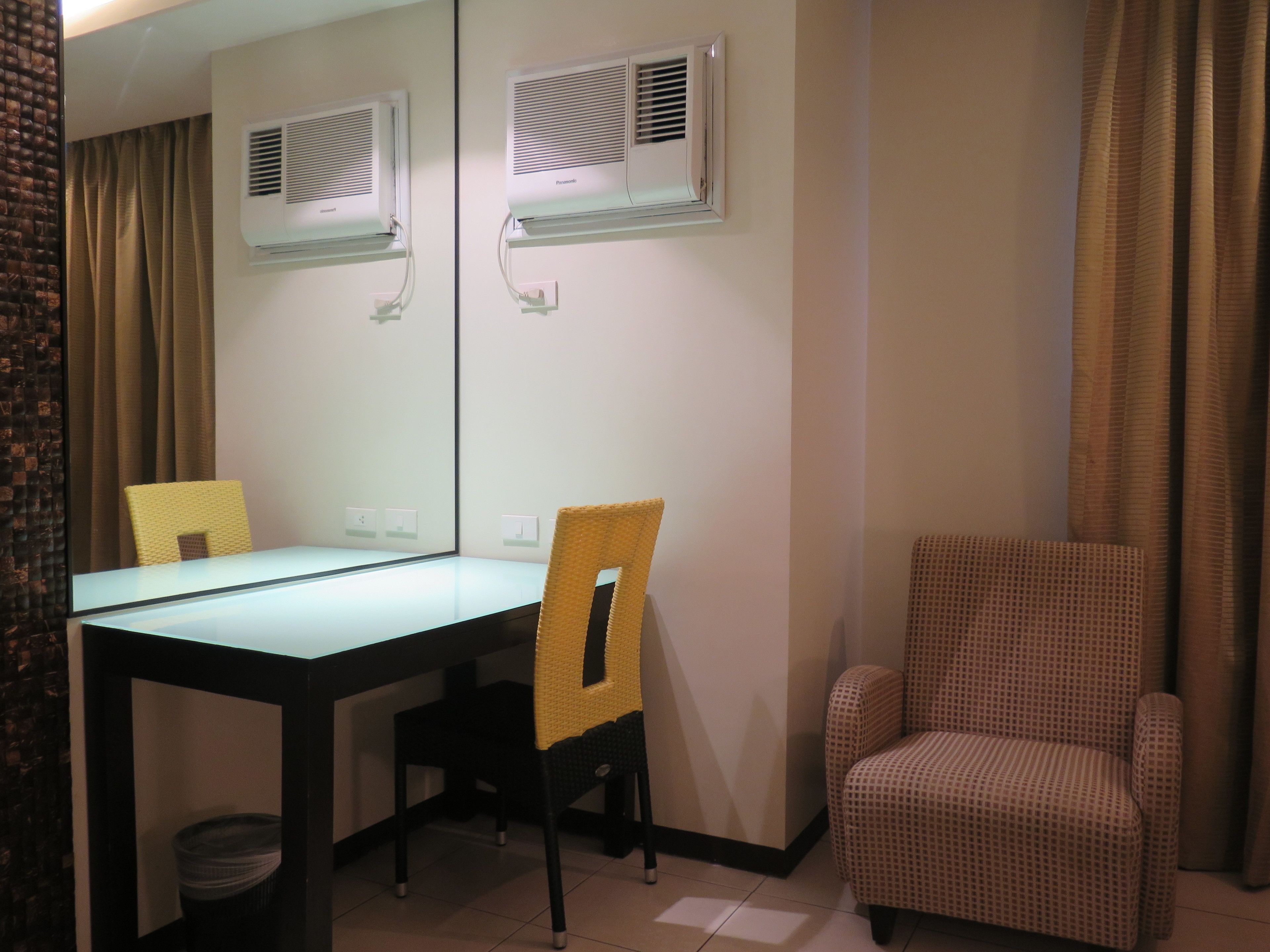 A hotel room with a dining table, two chairs, a sofa, and two wall-mounted air conditioning units.
