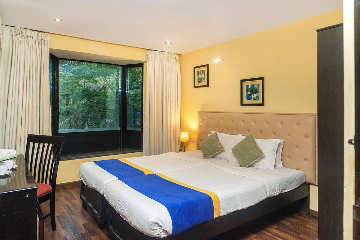 Una habitación de hotel con una cama, un escritorio y una silla. Hay una ventana con cortinas, una pared con dos cuadros enmarcados y una puerta.