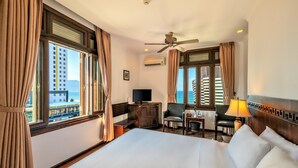 A hotel room with a large bed, a desk with a lamp, a chair, and a television. There are curtains on the windows, and an air conditioning unit is mounted on the wall. - Chu Hotel (Da Nang)