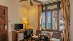 A hotel room with a wooden desk, a television, two chairs, a small table with a lamp, and a window with curtains. - Chu Hotel (Da Nang)