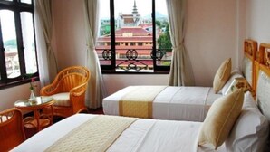 A hotel room with two beds, a small table, a wicker chair, and a view of a temple through large windows. - Areca Hotel (Hue)