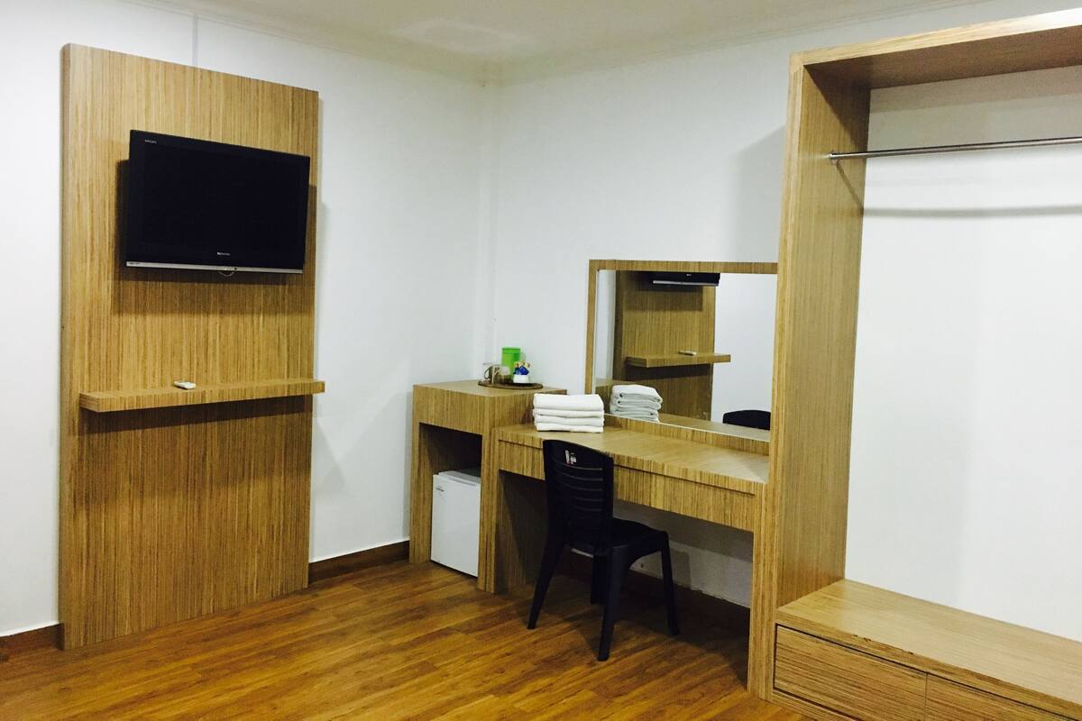 A compact room with a wooden desk, a chair, a TV mounted on the wall, a small fridge, and a mirror.