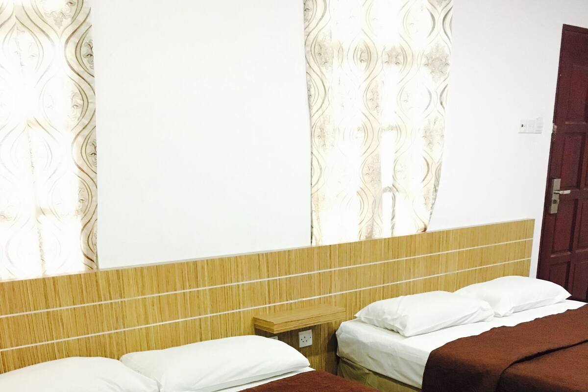 A hotel room with two beds, each with a brown bedspread and white pillows. There is a wooden nightstand between the beds, and the room has a wooden floor and a patterned curtain.
