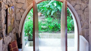 An interior with stone walls, wooden beams, a bench with a red cushion, and a view of greenery outside.