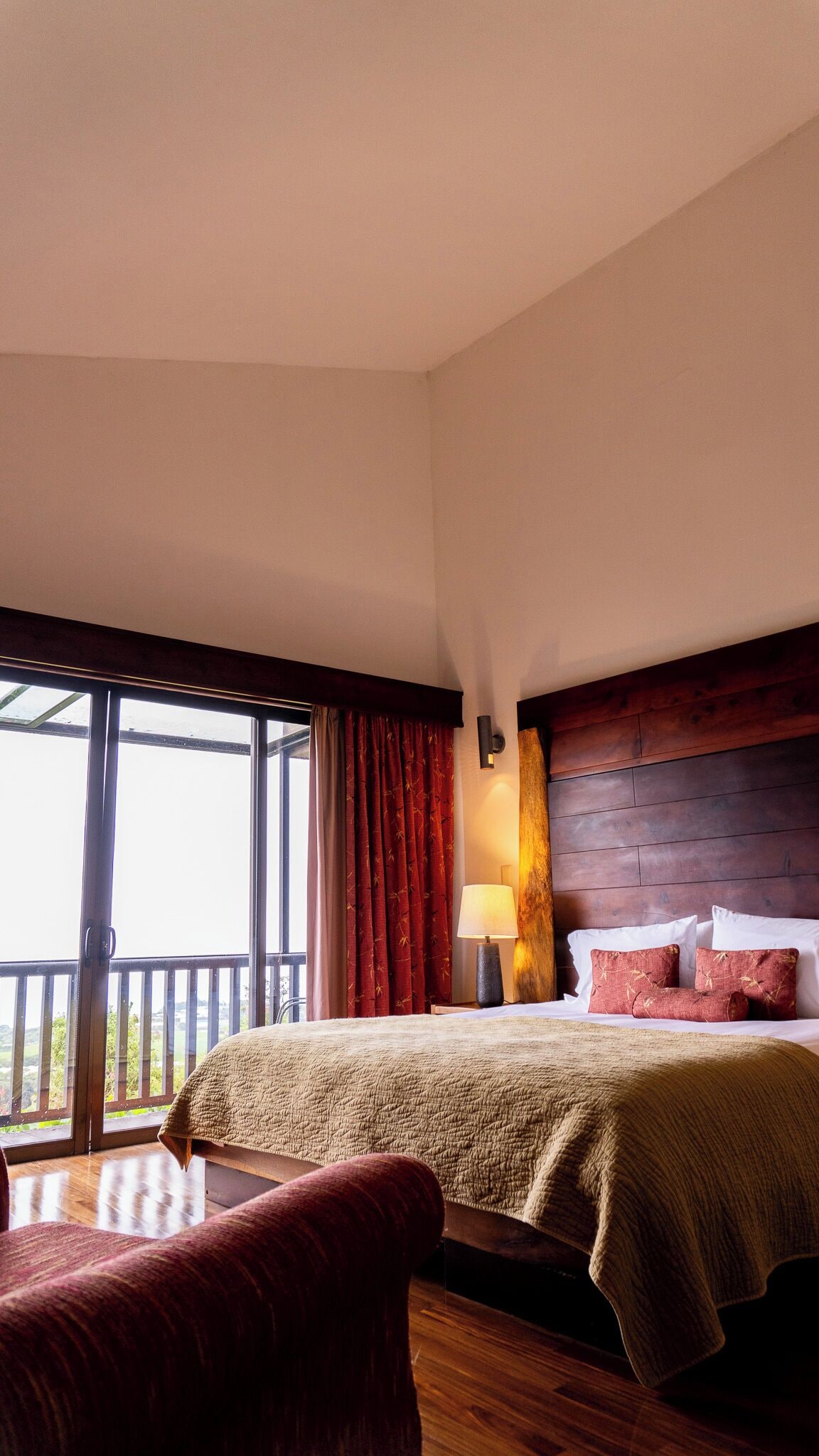 Una habitación de hotel con una cama grande, un cabecero de madera, una mesita de noche con lámpara y vistas amplias por una ventana.