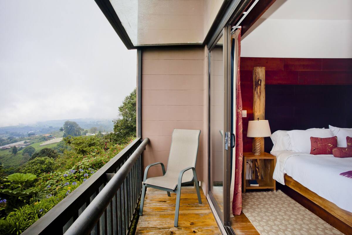 Un balcón con una silla, una cama con mesita de noche y vistas a un paisaje verde.